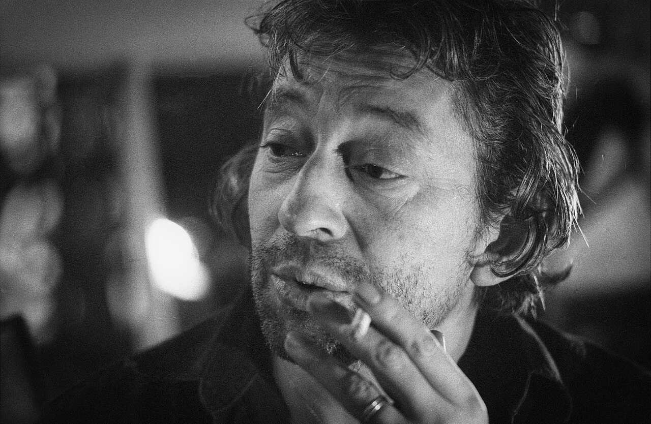 Foto von Gainsbourg mit langen Haaren und Dreitagebart. Er hält eine Zigarette in seiner linken Hand.